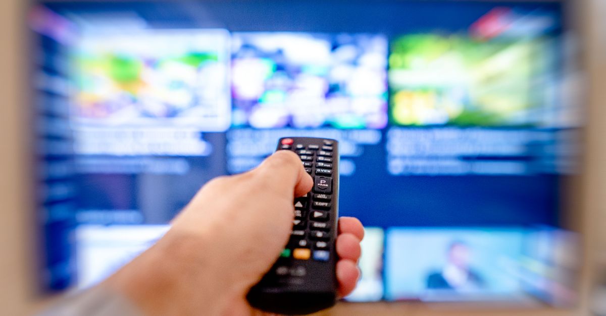 Hand holding remote control pointing at a blurred TV screen in a cozy indoor setting.